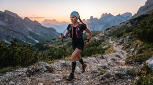 Female trail runner wearing a blue headband and hydration vest, running on a rocky mountain path at sunset.