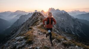 Trail runner in an orange jacket and black vest crosses a rocky mountain ridge at sunrise, bib 217 visible.