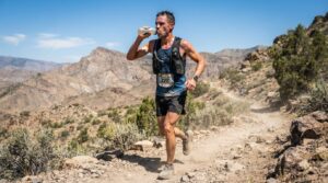 Male trail runner wearing bib 328 drinks water while running on a rocky desert trail with mountains in the background, sunlight brightening scene.